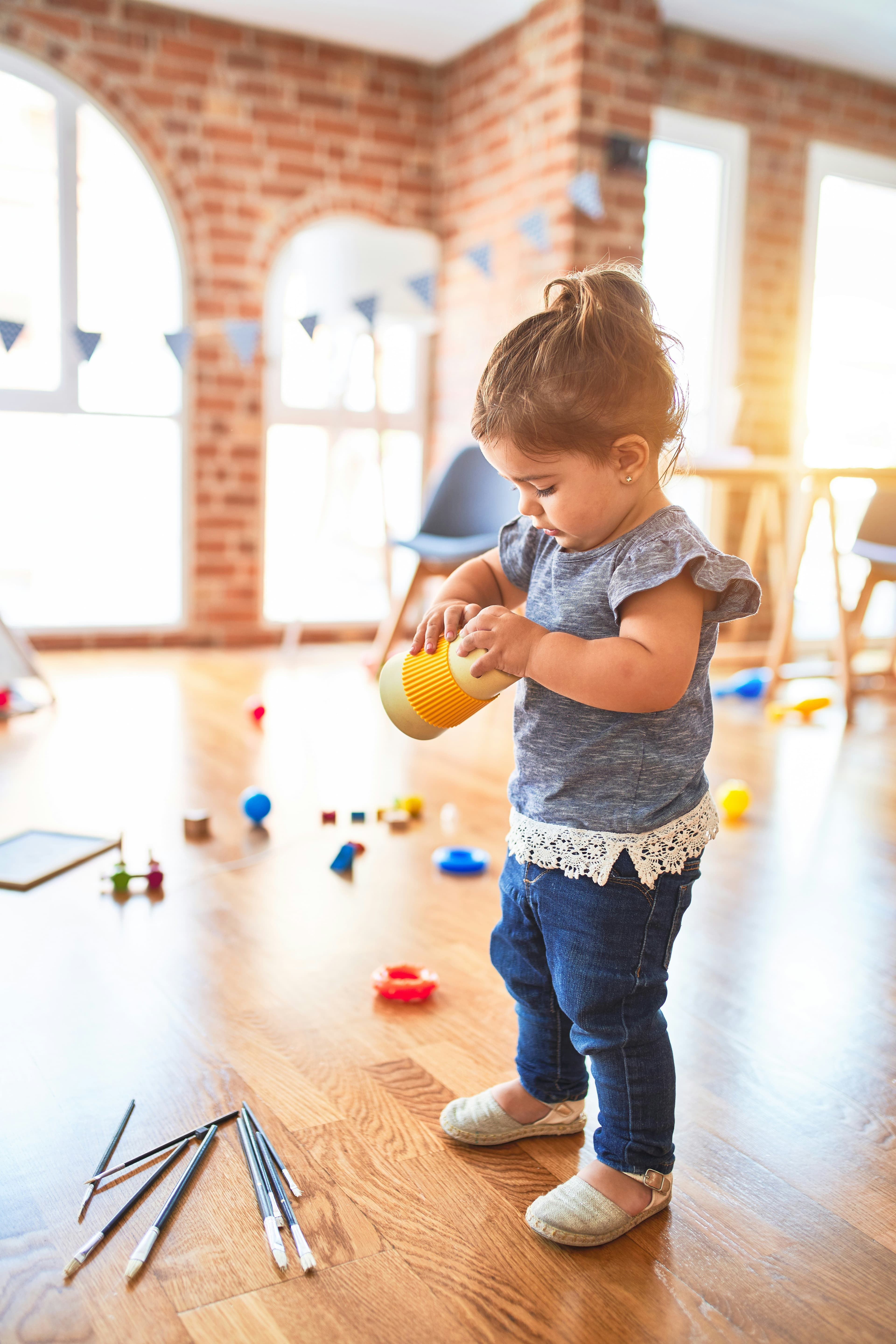 A young child plays with a yellow stacking toy in a bright, spacious classroom, surrounded by colourful educational toys and art supplies on the wooden floor.