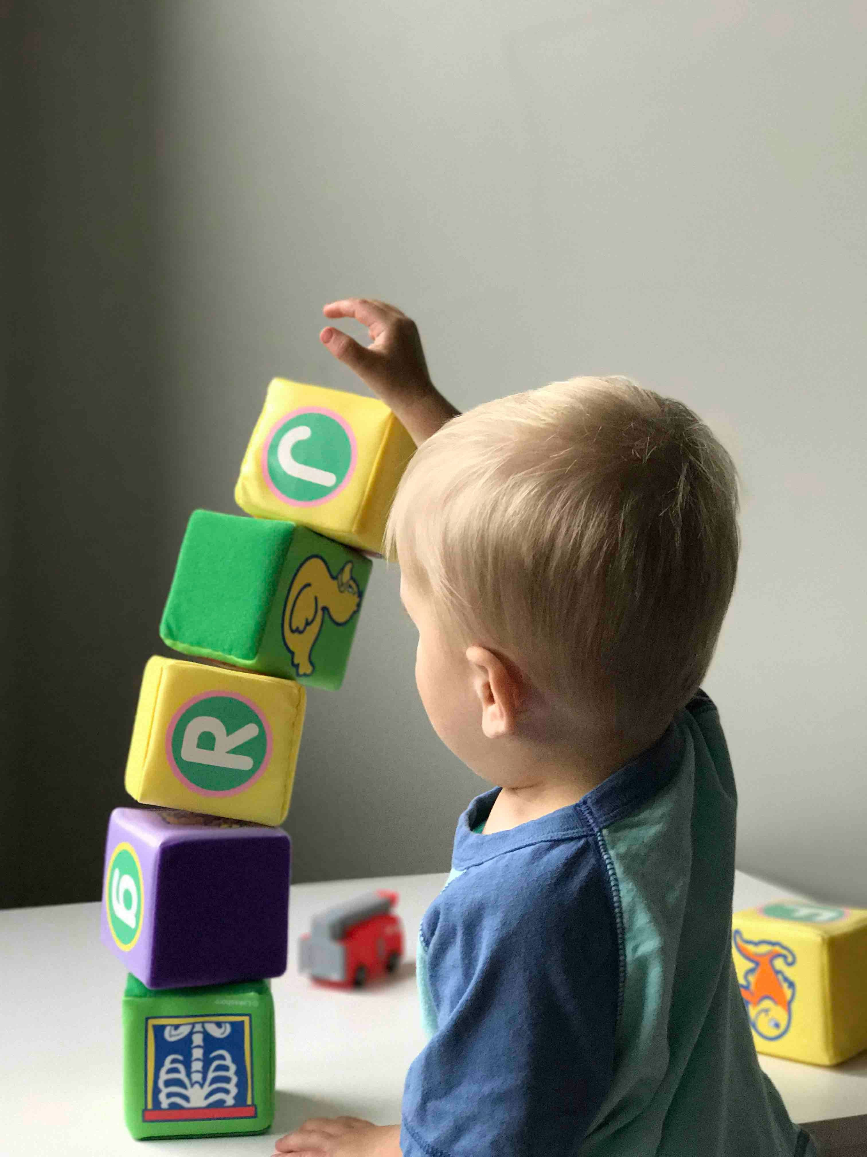 A young child stacks colourful alphabet blocks, carefully reaching to place a yellow block on top of the tower.